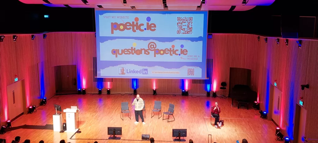 Kalle Ryan onstage at TU Dublin as MC and presenter at International Digital Marketing Week, Onscreen is a slide from his creative workshop that shows the web address for poetic.ie plus a QR code, an email address (questions@poetic.ie) and Kalle Ryan's LinkedIn profile with a QR code. 