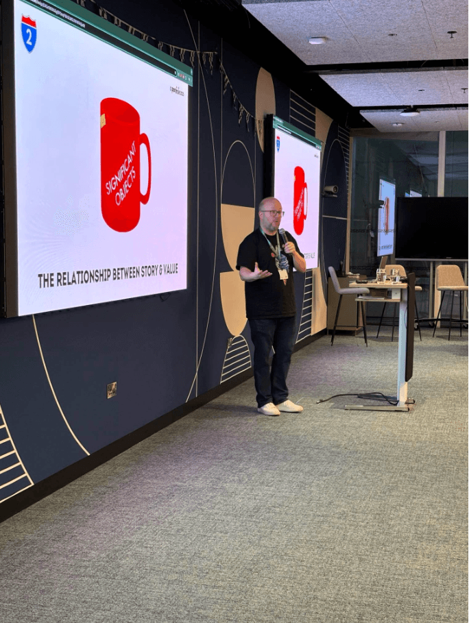 Kalle Ryan delivering a Storytelling Workshop at LinkedIn EMEA HQ in Dublin. Kalle is holding a mic in one hand and his other hand is directing the audience's attention to a slide with an image of a red mug with the caption "The relationship between story and value"